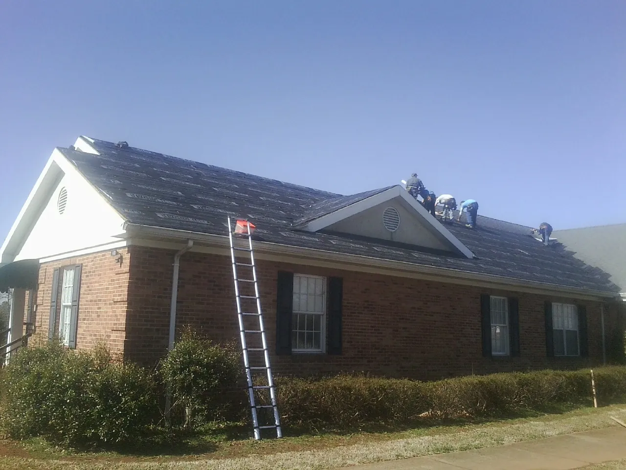 Crew tearing shingles off a brick ranch roof with a ladder set — Emergency Roof Repair work by our crew in Lake Mills